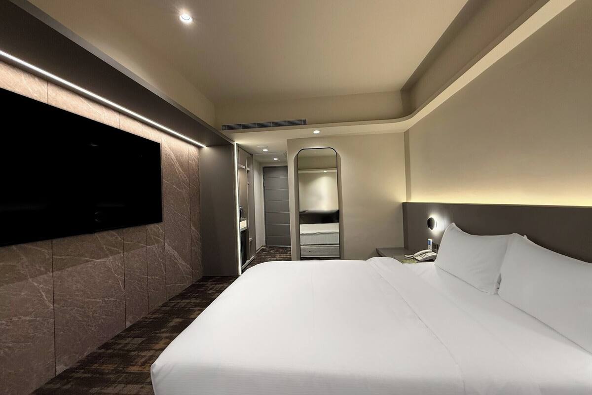 Una habitación de hotel moderna con una cama grande, un televisor de pantalla plana montado en la pared, una mesita de noche con lámpara y un vestidor.