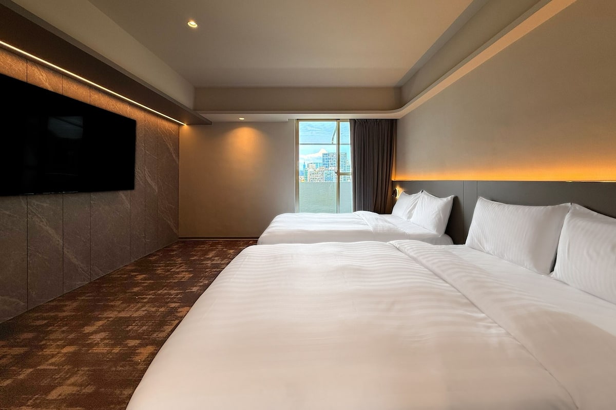 Habitación de hotel con dos camas, un televisor de pantalla plana en la pared y un amplio ventanal con vistas a la ciudad.