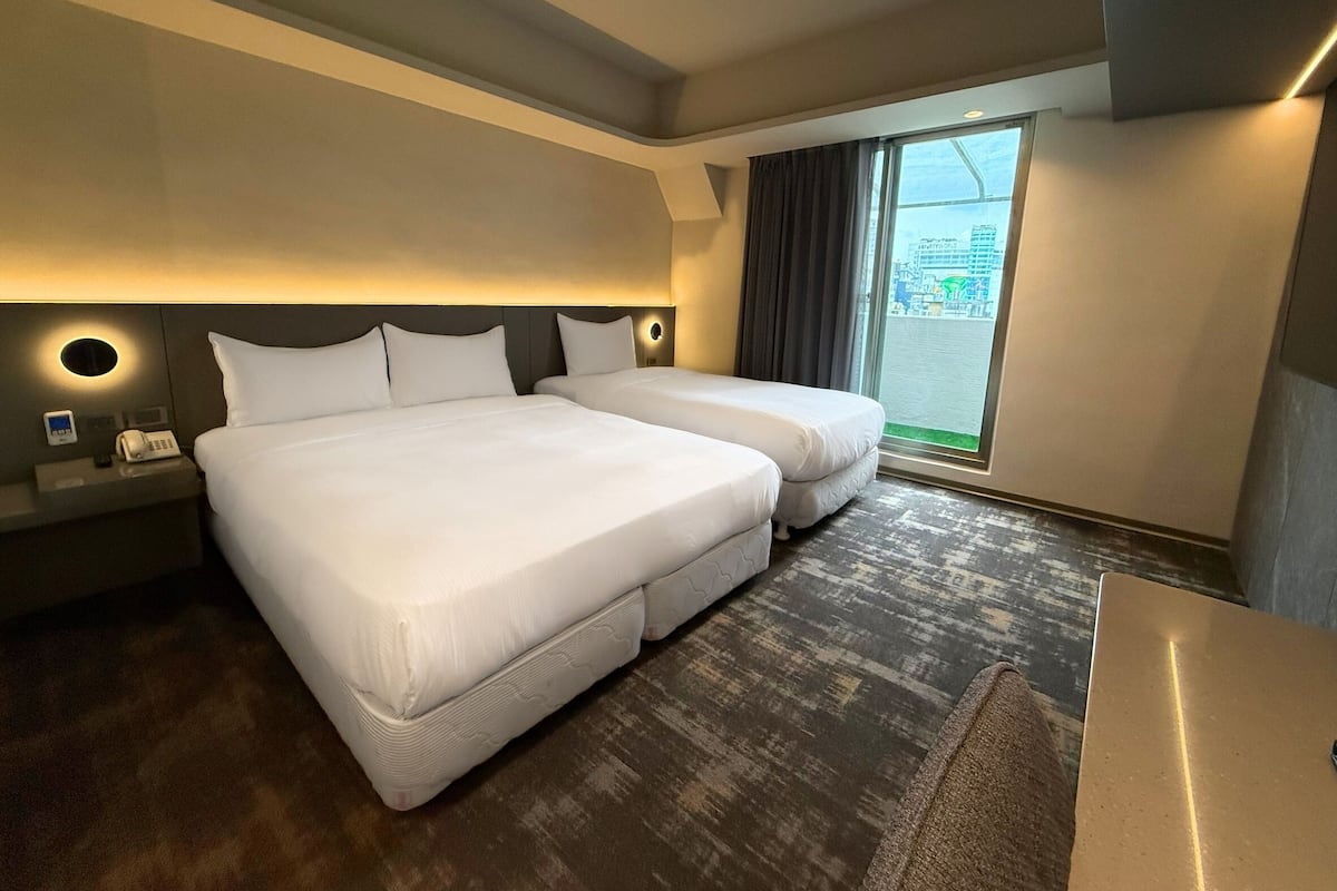 Habitación de hotel con dos camas, un escritorio, un teléfono y un ventanal con vistas a la ciudad.