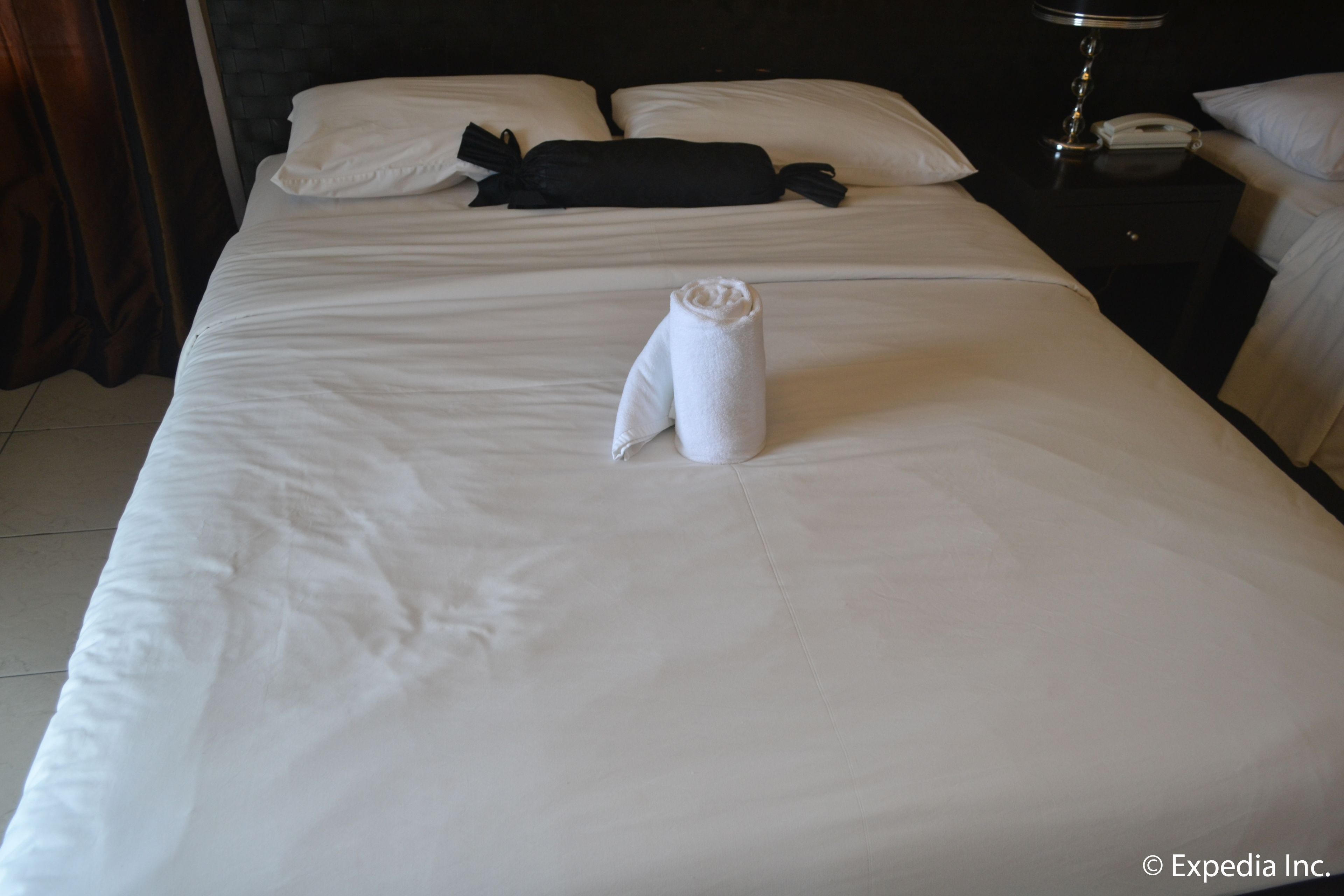 A neatly made bed with a rolled towel in the center, a black neck pillow, and a white pillow on the headboard.
