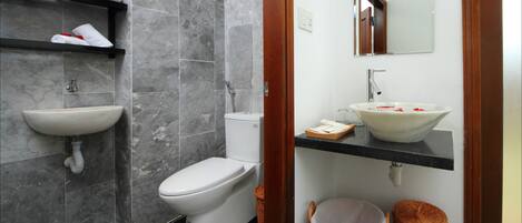 Ein Badezimmer mit Toilette, Waschbecken und Spiegel darüber. Unter dem Waschbecken befinden sich Regale mit Handtüchern und ein Korb auf dem Boden.