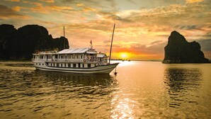 A boat with a covered deck and multiple windows, anchored near a coastline with rocky cliffs during sunset. - Halong Seasun Cruise (Bai Chay)