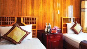 A room with wooden paneling, two beds with pillows, a nightstand with a bottle and fruit, and a window with a view of a body of water and cliffs. - Halong Seasun Cruise (Bai Chay)