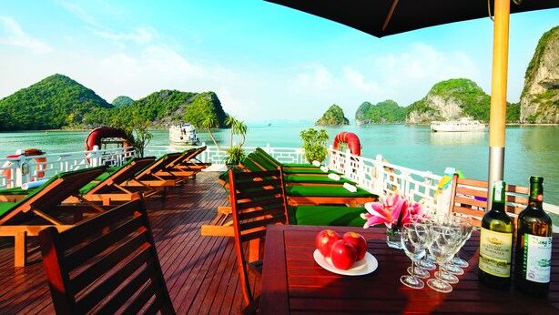 A wooden deck with lounge chairs and tables, overlooking a calm sea with boats and lush green islands. - Halong Seasun Cruise (Bai Chay)