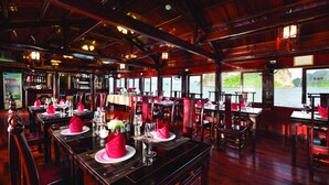 A restaurant with wooden interiors, tables set with red napkins and white plates, and a view of the water outside. - Halong Seasun Cruise (Bai Chay)