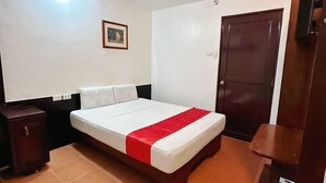 Hotelkamer met een bed, een houten nachtkastje, een klein kastje, een deur en een lamp aan het plafond.