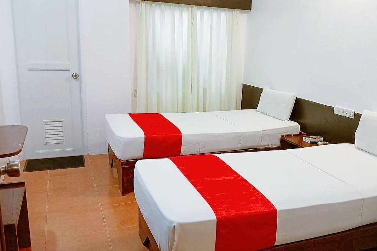 A hotel room with two beds, each with a red and white bedspread, a wooden headboard, a small table, and a window with white curtains.
