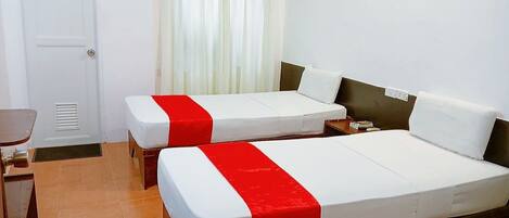 A hotel room with two beds, each with a red and white bedspread, a wooden headboard, a small table, and a window with white curtains.