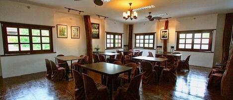 Un amplio comedor con múltiples mesas y sillas, ventanas grandes, ventiladores de techo y obras de arte en las paredes.