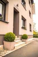 Entrada de un hotel con un letrero que dice “HOTEL” y un banderín sobre la puerta. Hay escalones que conducen a la entrada, y dos macetas a cada lado.