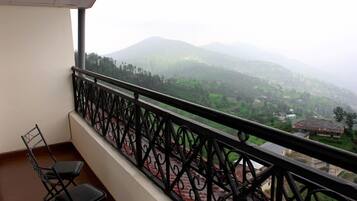 A balcony with a wrought iron railing, two black chairs, and a view of green hills and buildings in the distance.