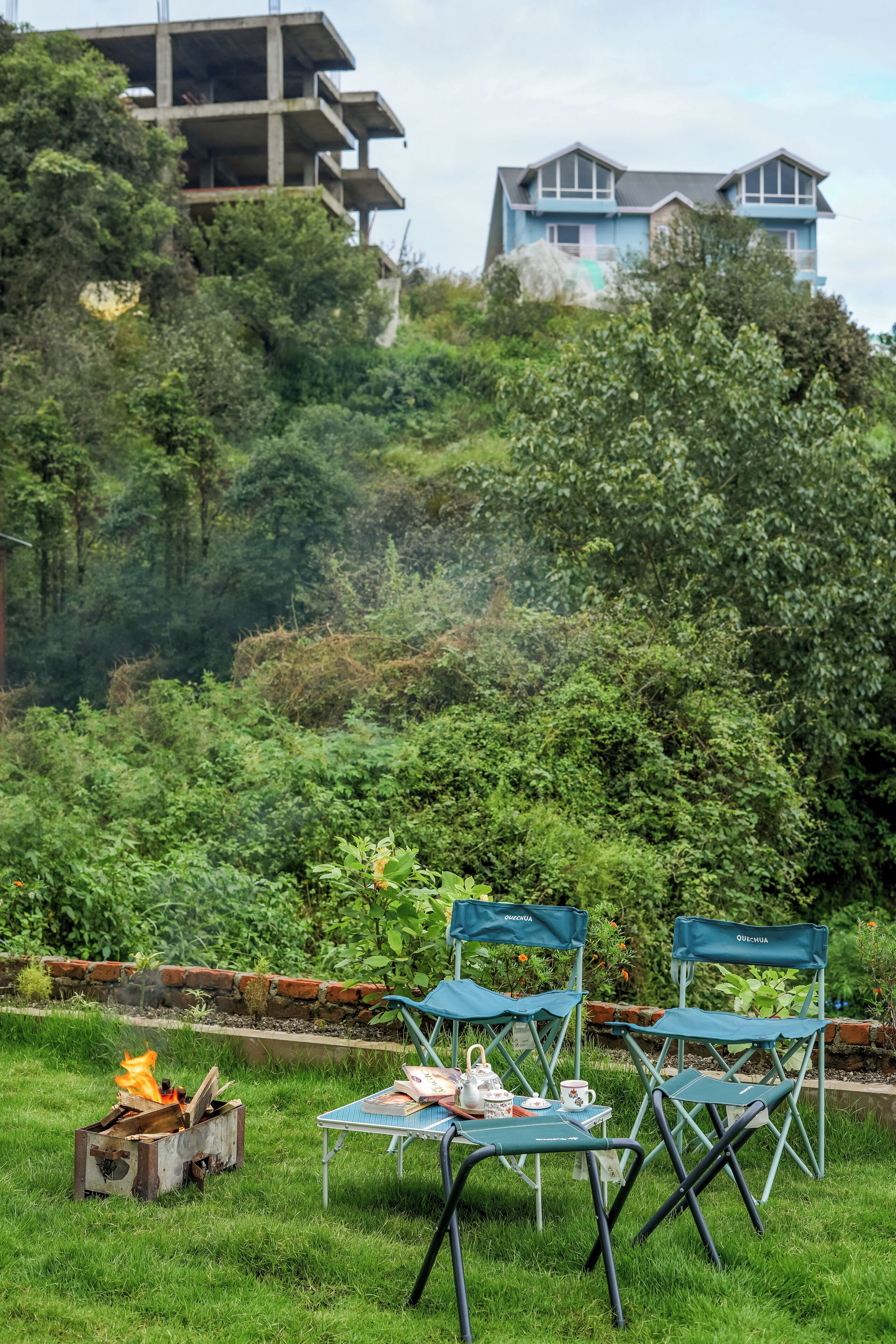 Un área de estar al aire libre con una chimenea, sillas plegables verdes y una mesa con libros y un calentador de agua.