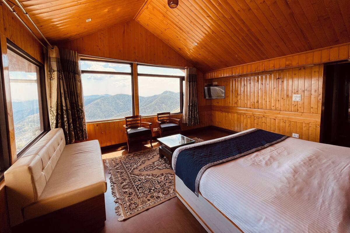 A cozy room with a bed, a sofa, a table, and a TV. Large windows offer a view of the mountains.
