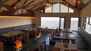 Een restaurant met houten tafels en stoelen, een hoog plafond met zichtbare balken en grote ramen met uitzicht op de bergen.