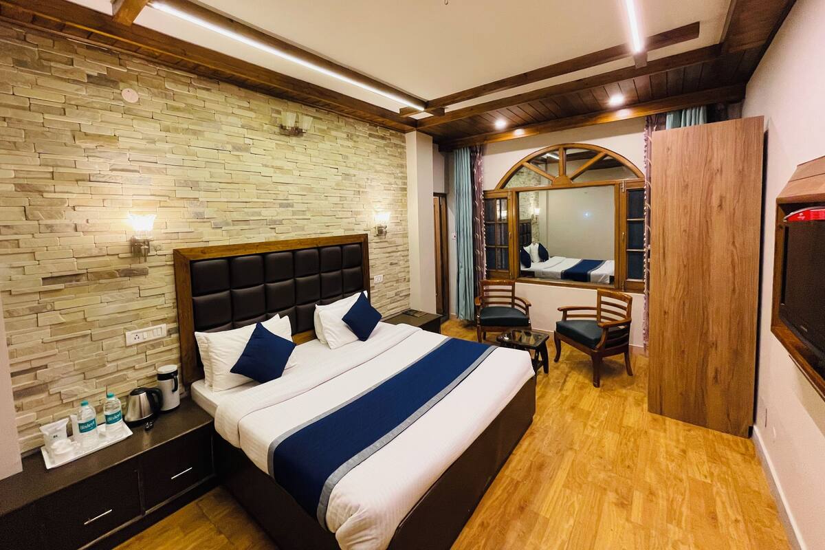 A bedroom with a stone wall, a bed with a headboard, two chairs, a wooden wardrobe, and a mirror.