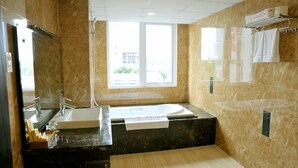 A modern bathroom with a large bathtub, a sink, and a shower. There is a window providing natural light and a view of the outside. - International Hotel (Can Tho)