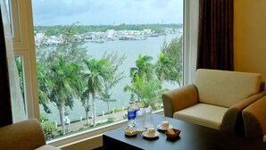 A room with a view of a body of water, a sofa, a table with a tray of items, and a window with curtains. - International Hotel (Can Tho)