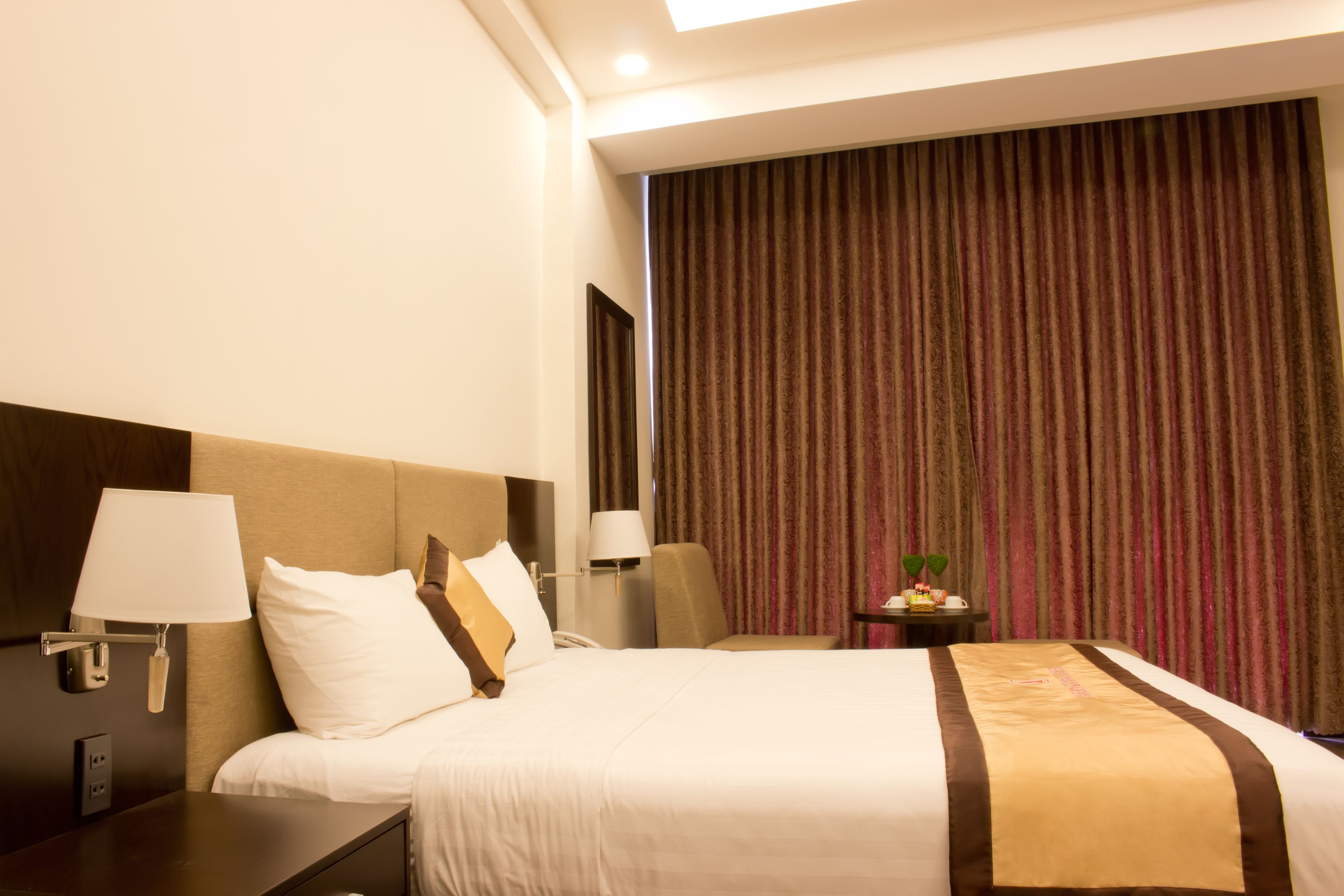 A hotel room with a large bed, a nightstand, a lamp, a mirror, a small table with a plant, and a curtain.
