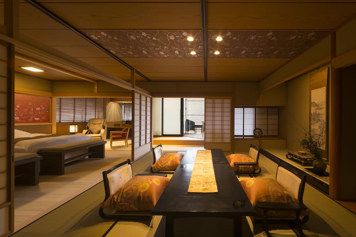 A traditional Japanese room with tatami flooring, a low table with cushions, a futon bed, sliding doors, and a view of a garden.