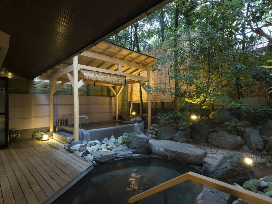 A traditional Japanese garden with a wooden deck, a hot spring, and a covered seating area.