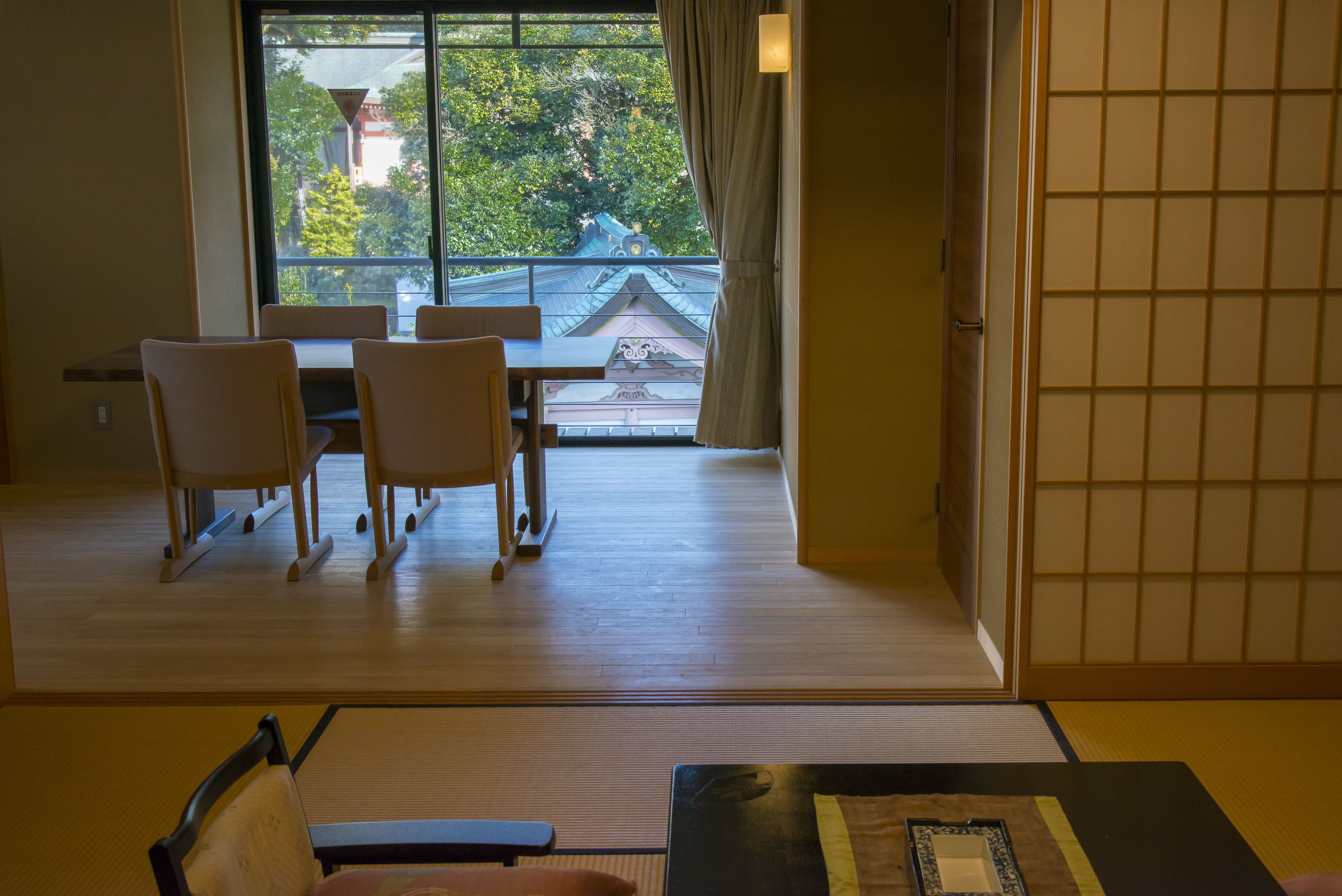 A traditional Japanese room with tatami flooring, a low table with a cushion, and a view of a traditional building outside.