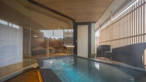 Un área interior de piscina moderna con paredes de vidrio transparente, suelo de madera y zona de estar con sillas.