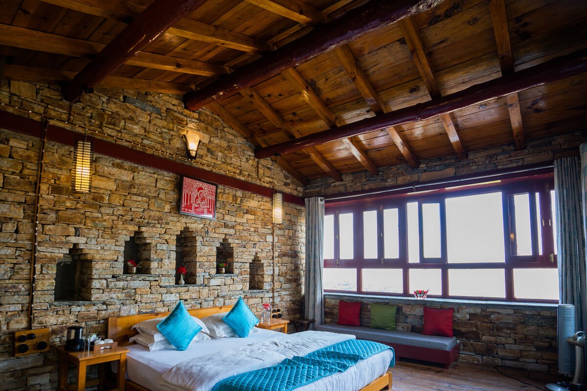 Ein rustikales Zimmer mit einer Steinwand, Holzbalken, einem Bett mit blauen Kissen, einem Fenster mit Rollläden und einem Steinkamin.