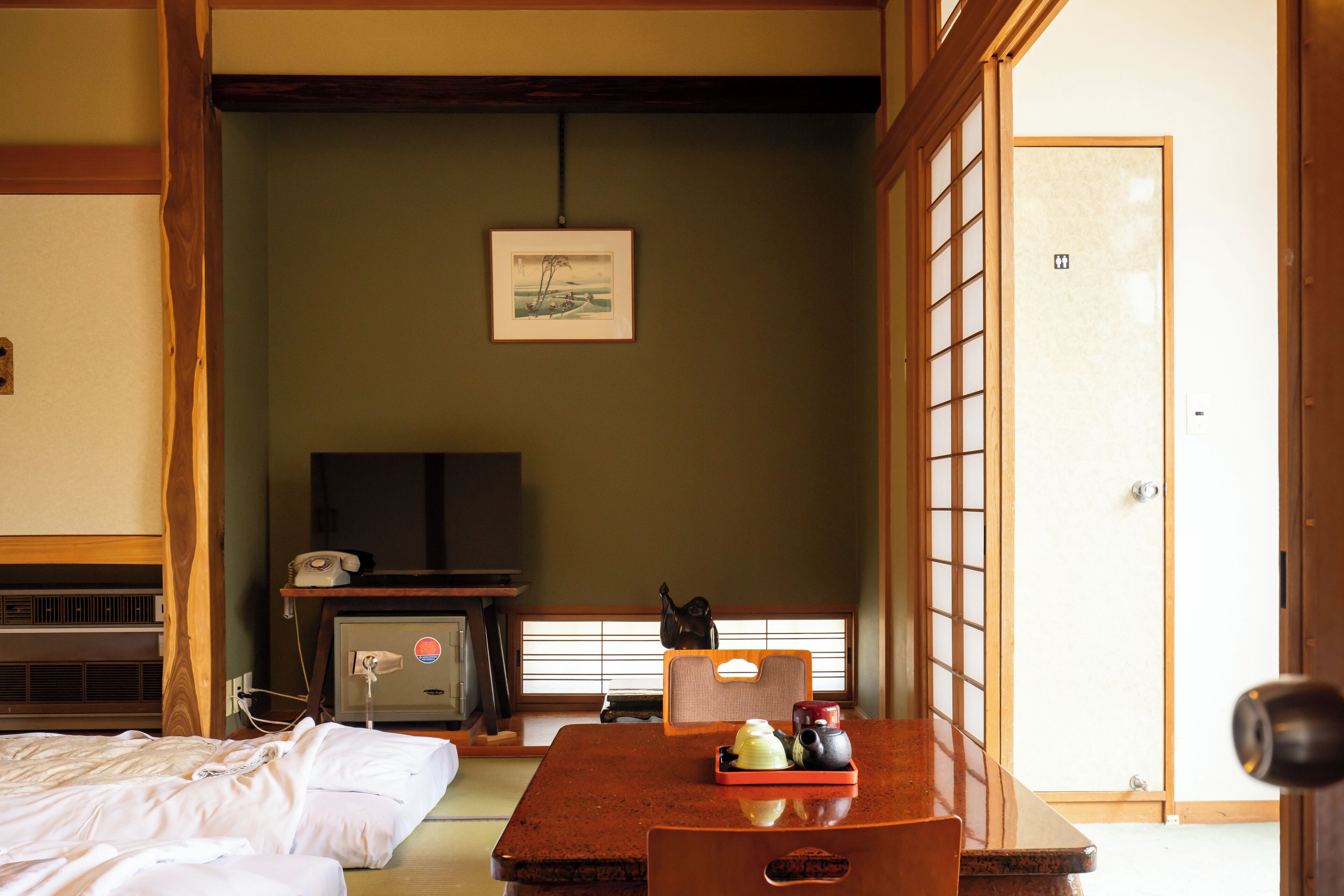 A traditional Japanese room with tatami flooring, a low table with a tray of tea utensils, a television on a cabinet, and a sliding door leading to another room.