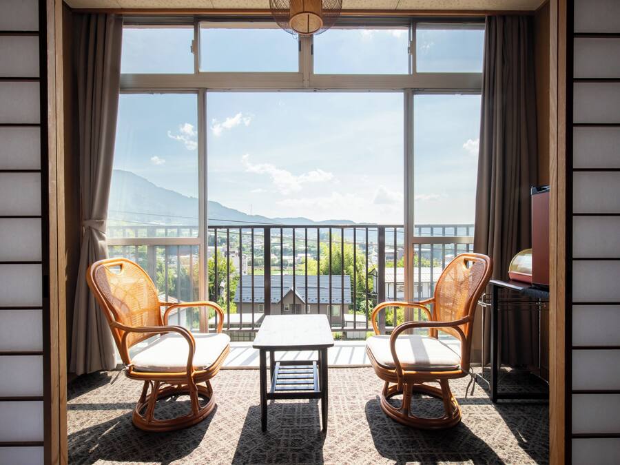 A balcony with two wicker chairs and a small table, overlooking a scenic view of mountains and a bridge.