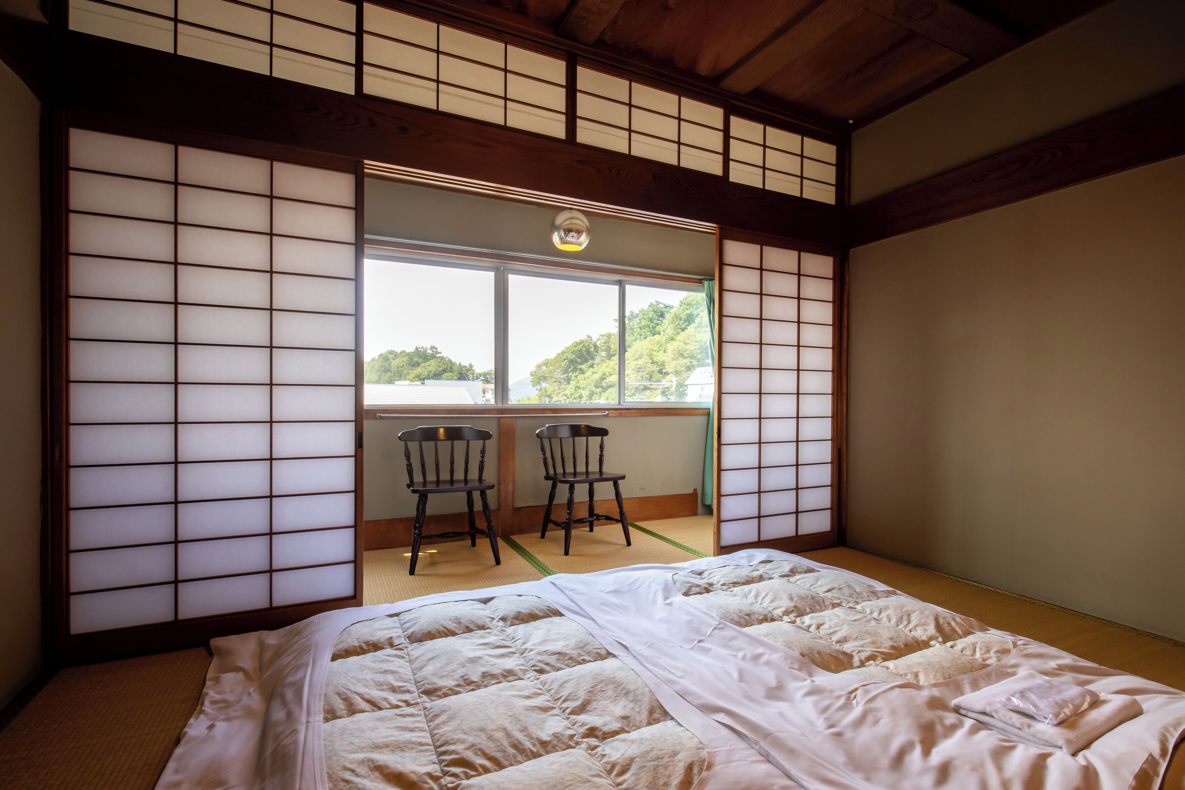 A traditional Japanese room with tatami flooring, a futon bed, sliding shoji doors, and a view of greenery outside.