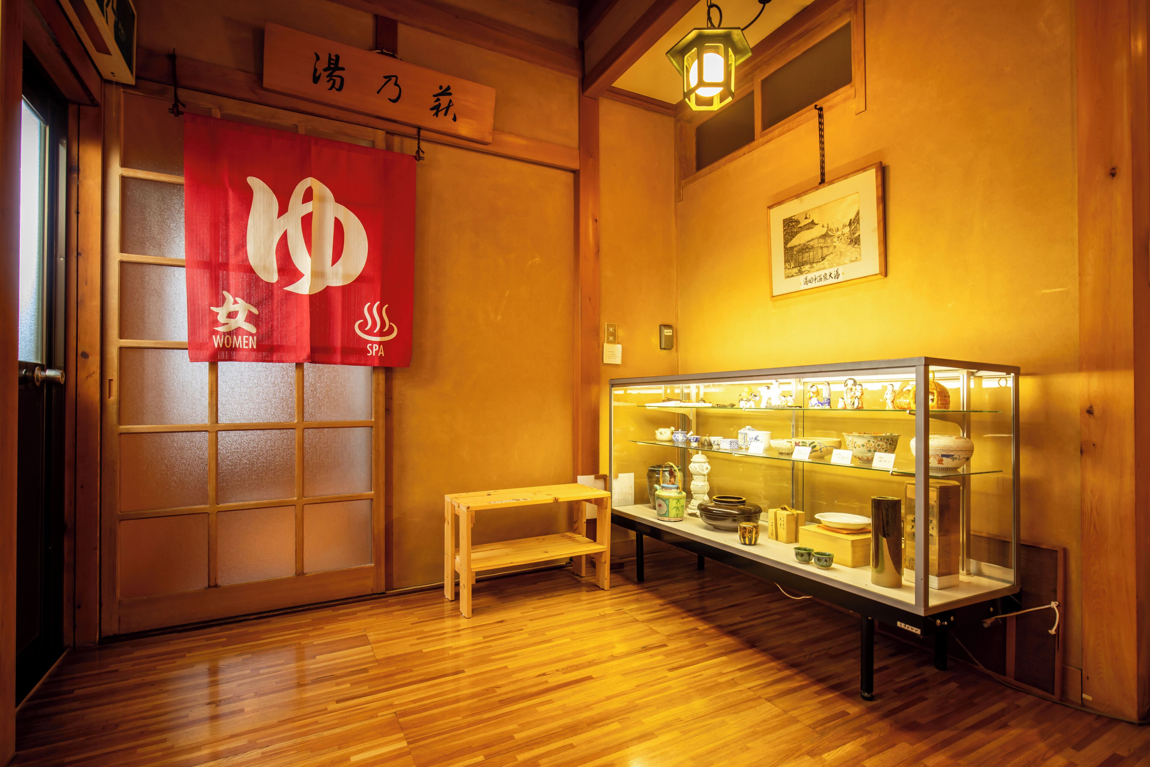 Une pièce de style japonais traditionnel, avec un plancher en bois, une vitrine contenant des objets en céramique et une bannière rouge ornée de caractères blancs.
