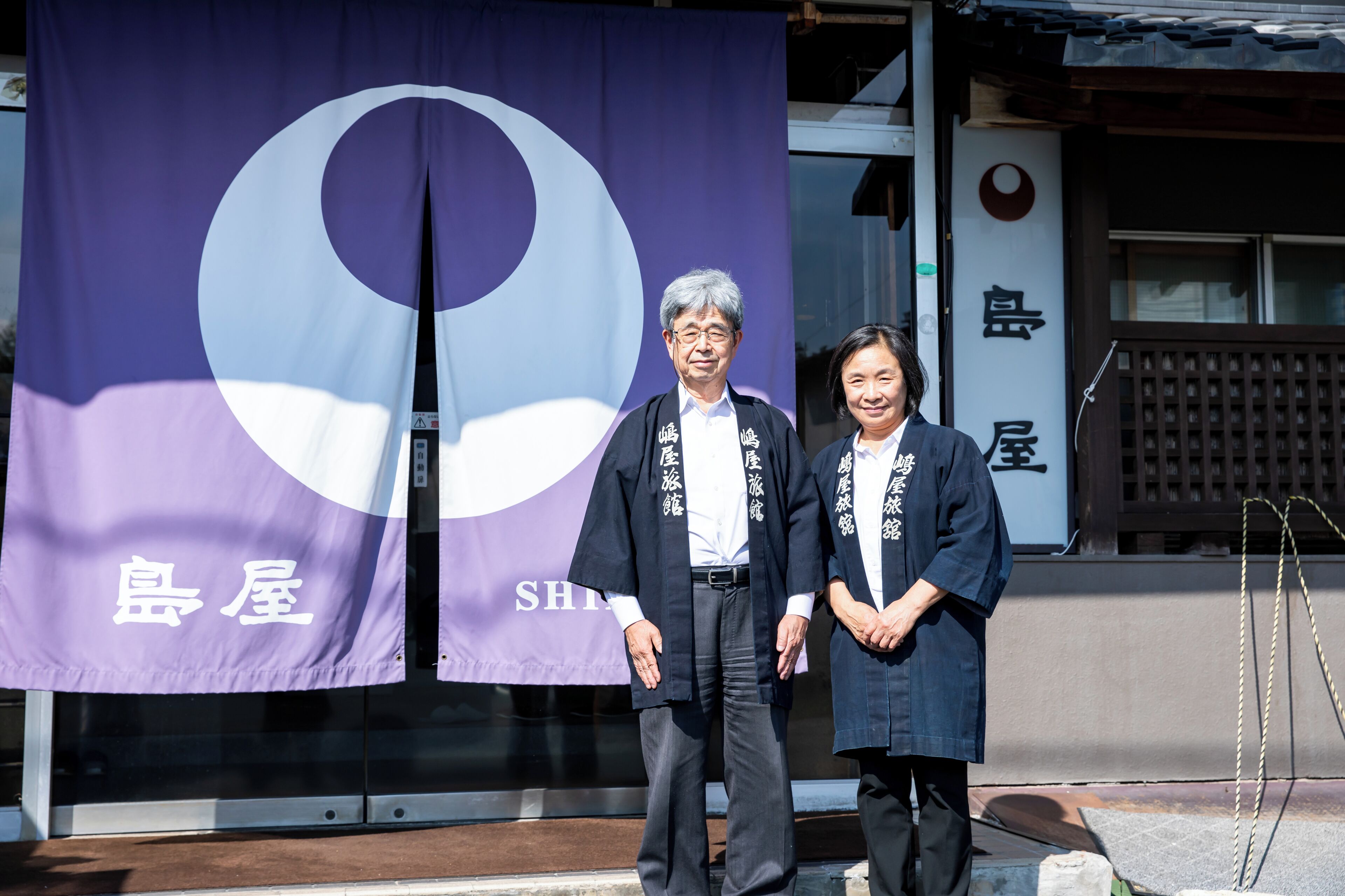 Dos personas de pie frente a un edificio con un cartel morado grande que tiene un círculo blanco y caracteres japoneses.