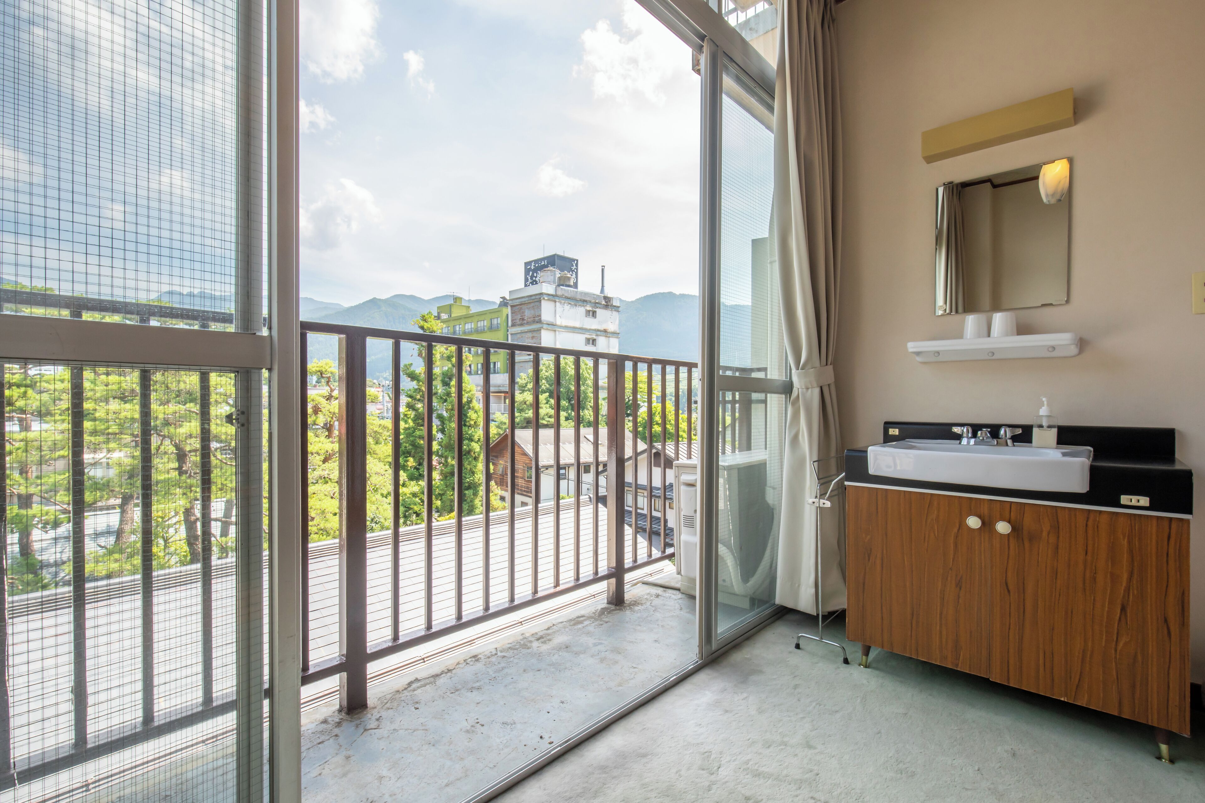 Un balcón con barandilla metálica, un tocador de madera con espejo y lavabo, y vistas a edificios y árboles.