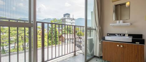 Un balcón con barandilla metálica, un tocador de madera con espejo y lavabo, y vistas a edificios y árboles.