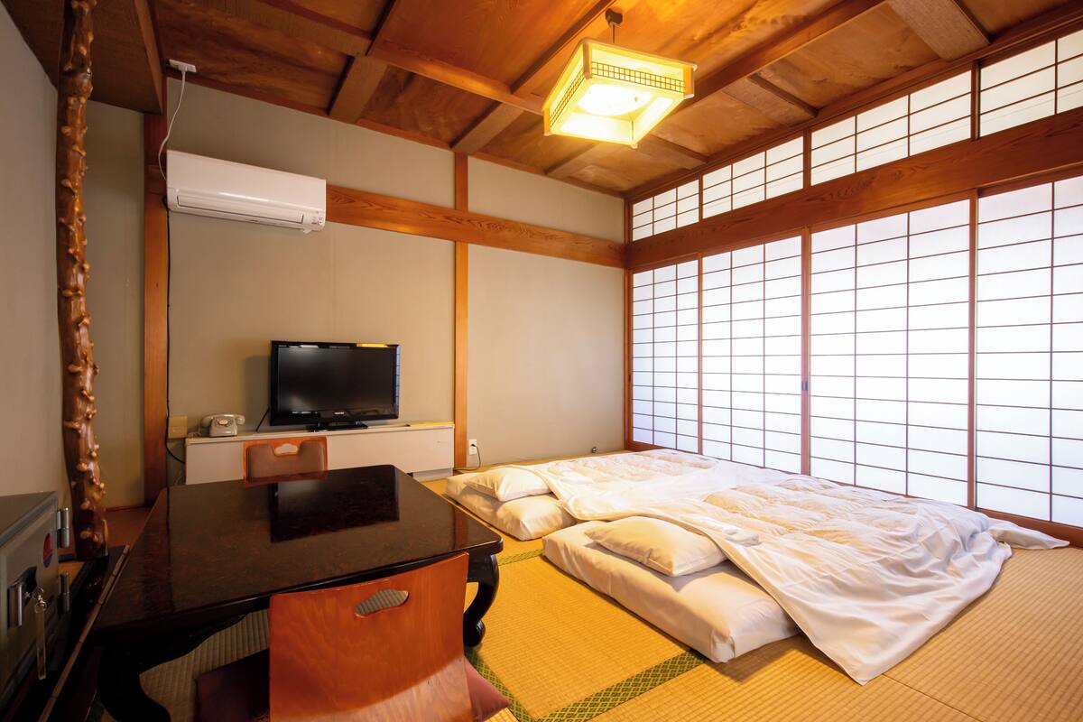 A traditional Japanese-style room with tatami flooring, a bed with white bedding, a television on a wooden table, and a shoji screen window.