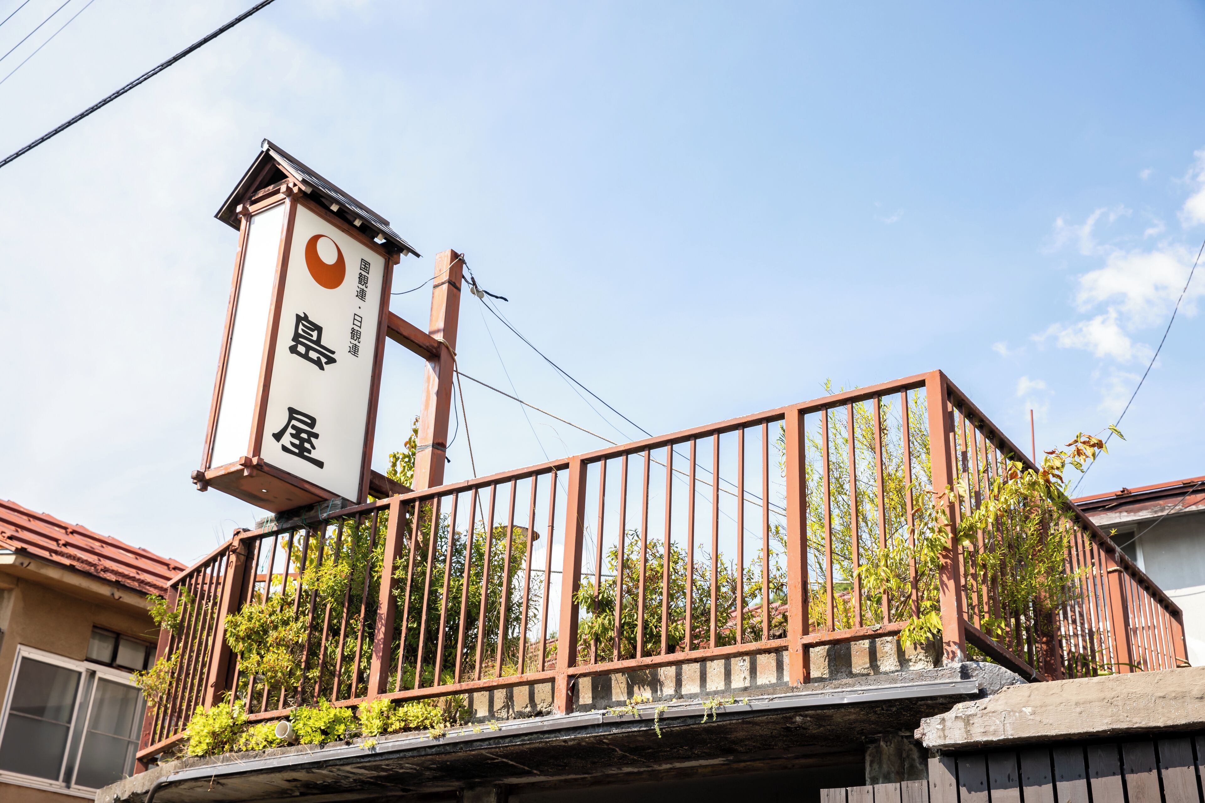 A traditional Japanese building with a signboard, a metal fence, and greenery.