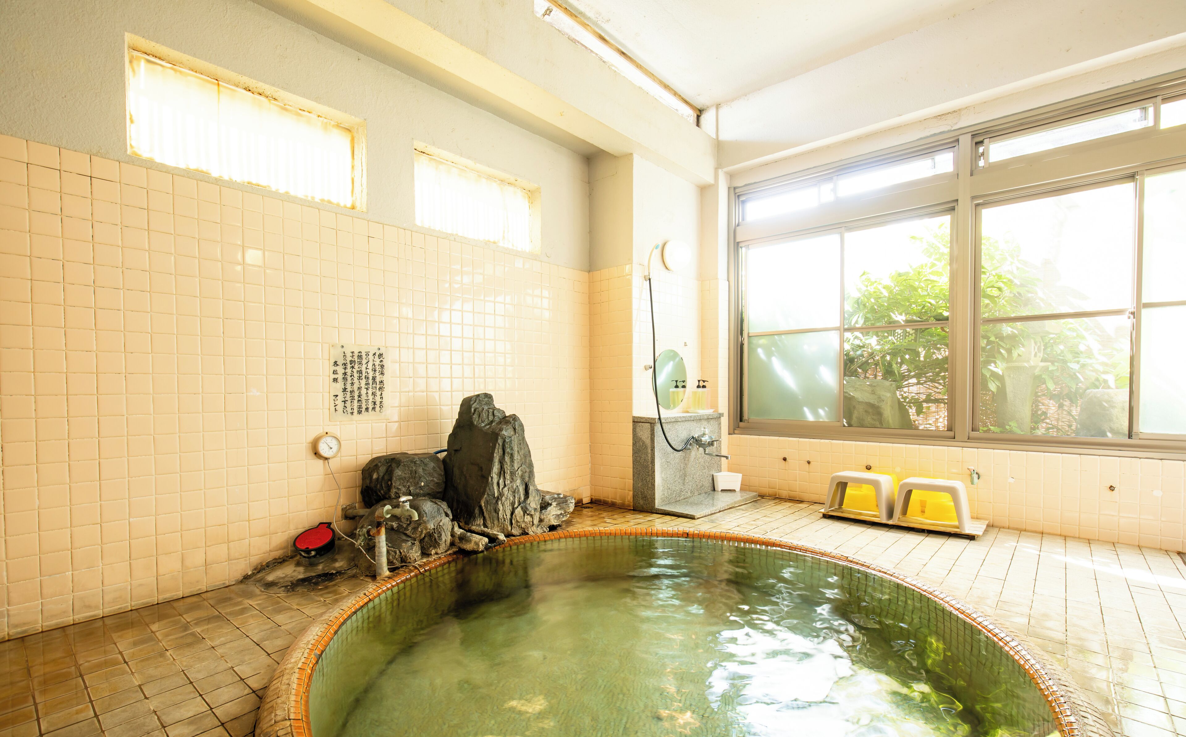 A traditional Japanese onsen with a stone bath, tiled walls, and a view of greenery outside.