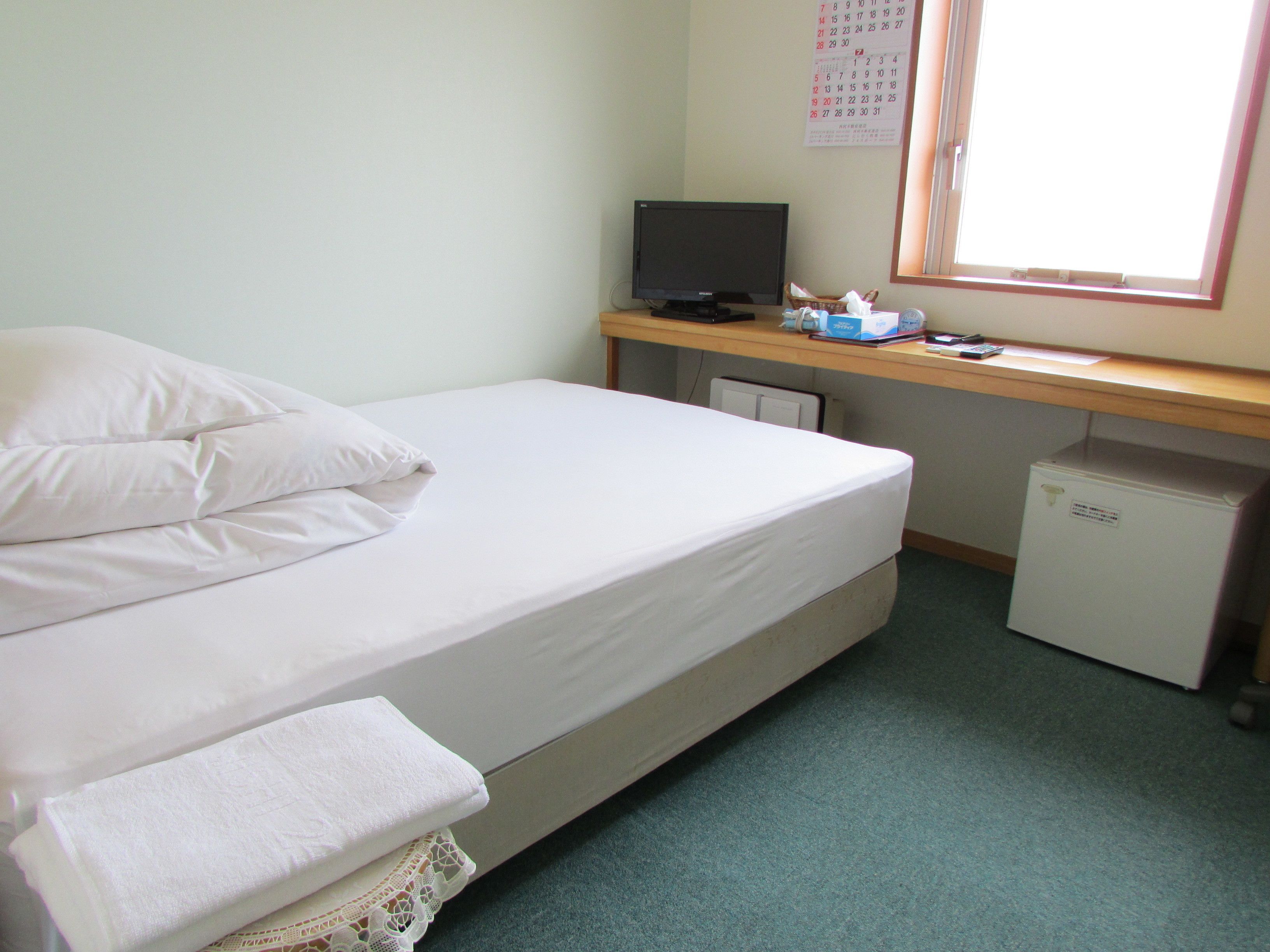 A single-bed room with a desk, a computer, a small fridge, and a window with a calendar.