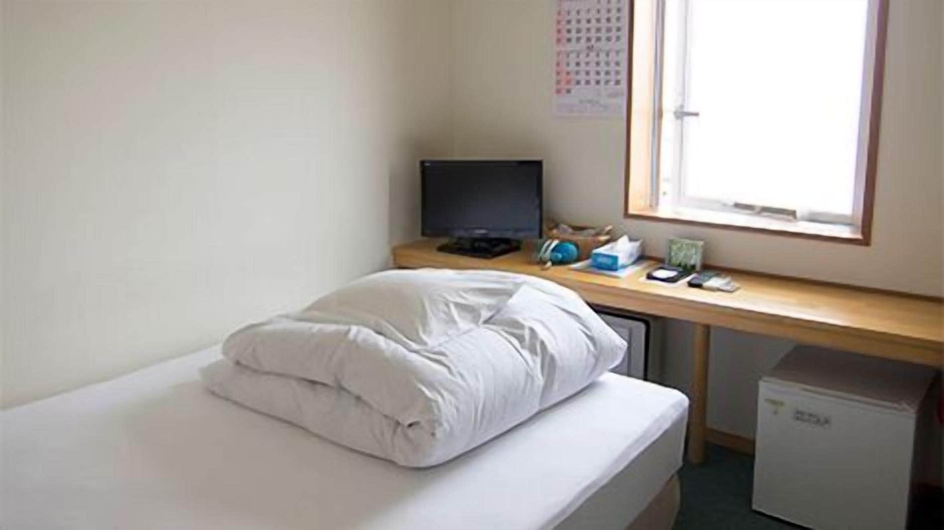 A small, neatly made bed with white bedding in a room with a wooden desk and a computer monitor.
