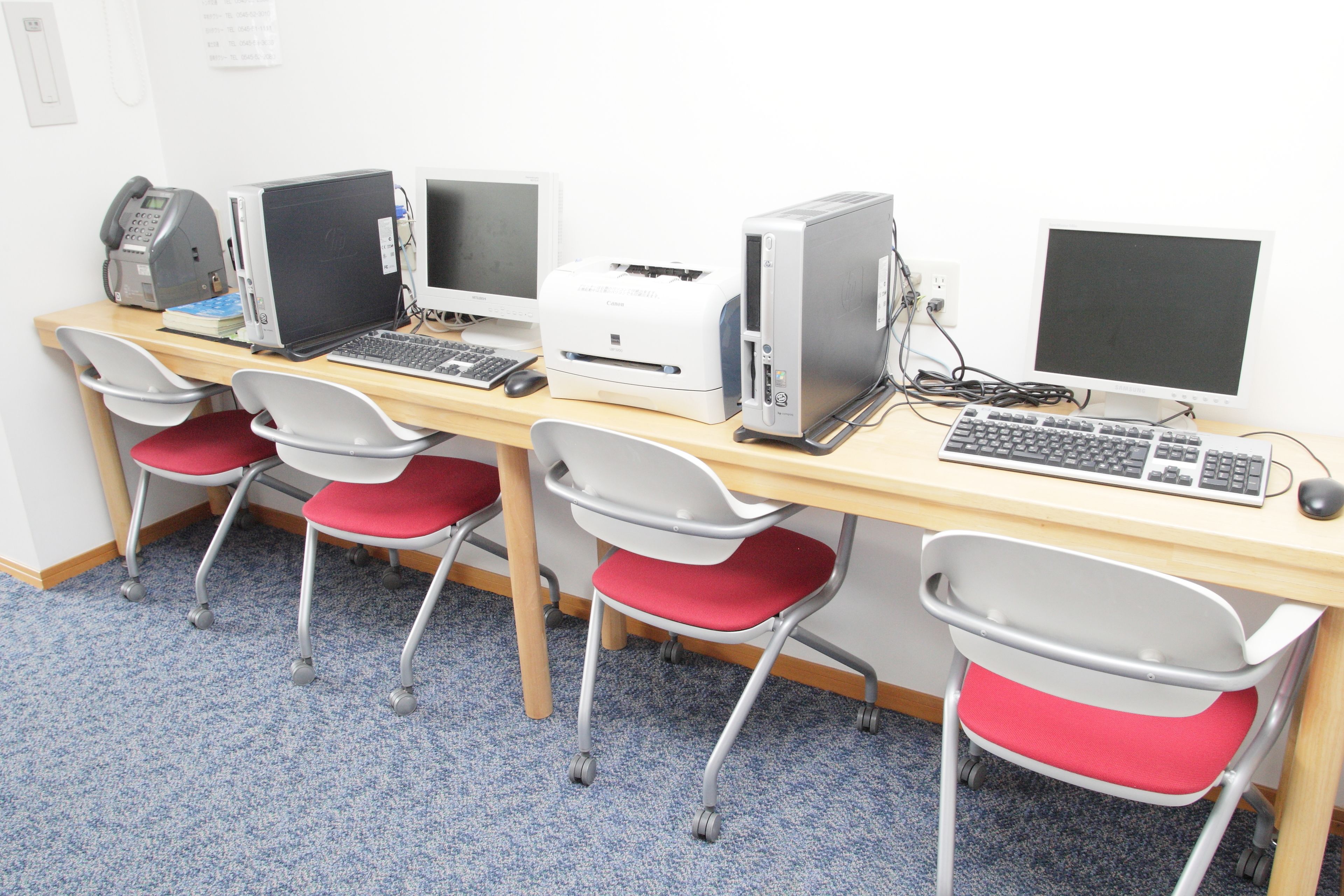 Un laboratorio de computación con cuatro escritorios, cada uno con una computadora de escritorio, teclado y ratón. Hay cuatro sillas con cojines rojos.