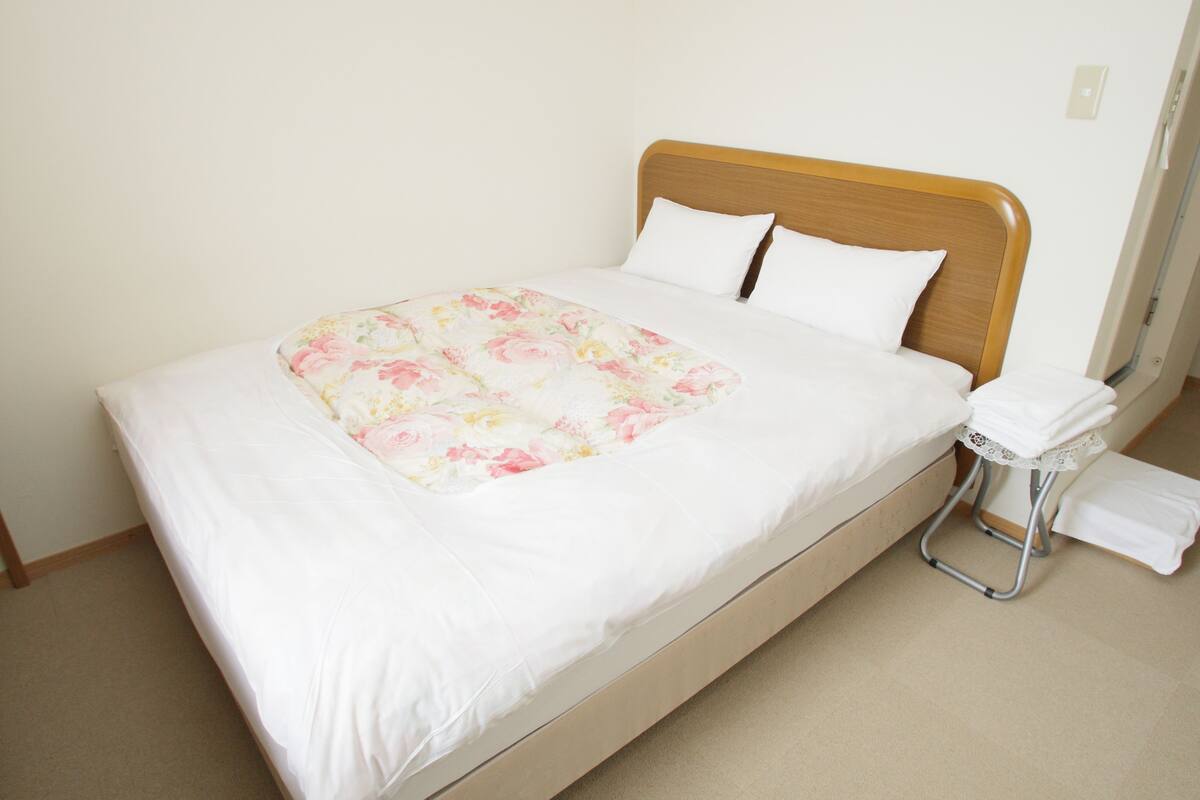 A neatly made bed with a floral bedspread and two pillows, a bedside table with a towel, and a plain wall with a light switch.