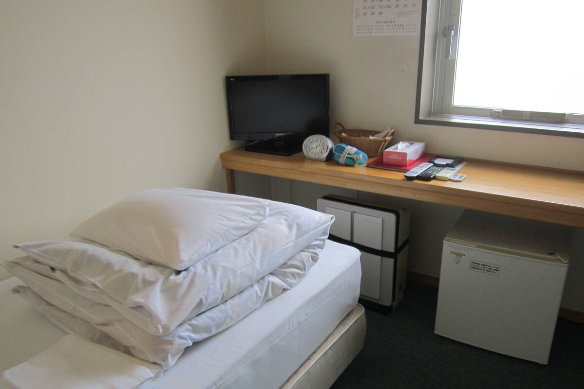 A single bed with white bedding, a small desk with a computer, a television, and a refrigerator.
