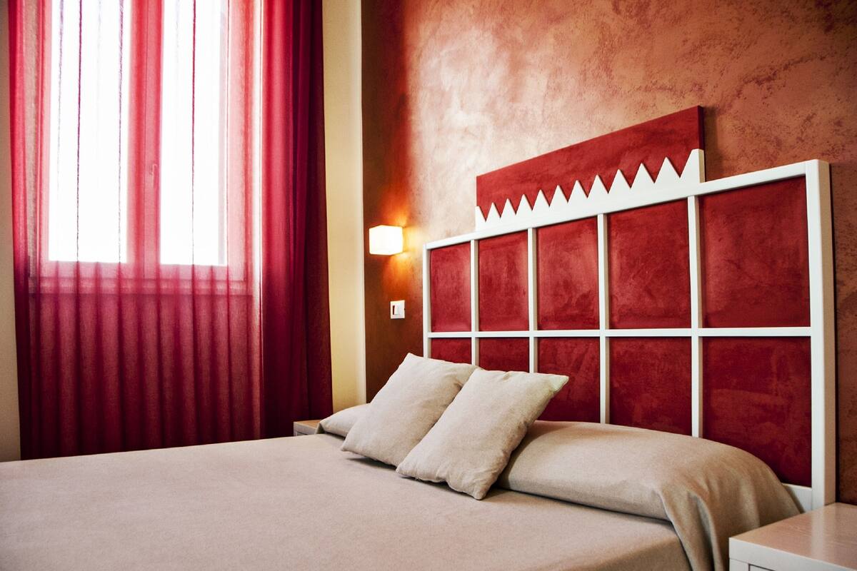 Una camera d'albergo con un letto grande, tende rosse, una testiera rossa e una lampada fissata al muro.