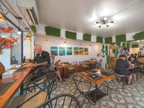 L’intérieur d’un restaurant, avec de nombreuses tables et chaises, des œuvres d’art accrochées aux murs et un ventilateur de plafond.