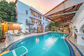 A swimming pool with lounge chairs and umbrellas, surrounded by a building with a covered patio area. - Mali Namphu Hotel (Vientiane)