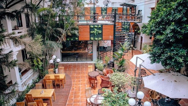 Un patio con varias mesas y sillas de madera, rodeado de abundante vegetación y una escalera de caracol que conduce a un nivel superior.