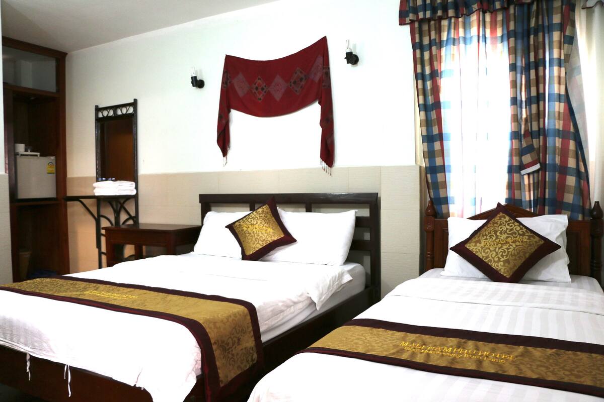 Habitación de hotel con dos camas, cada una con colcha blanca y detalles dorados, cabeceros de madera, una chalina roja colgada en la pared y una ventana con cortinas estampadas.