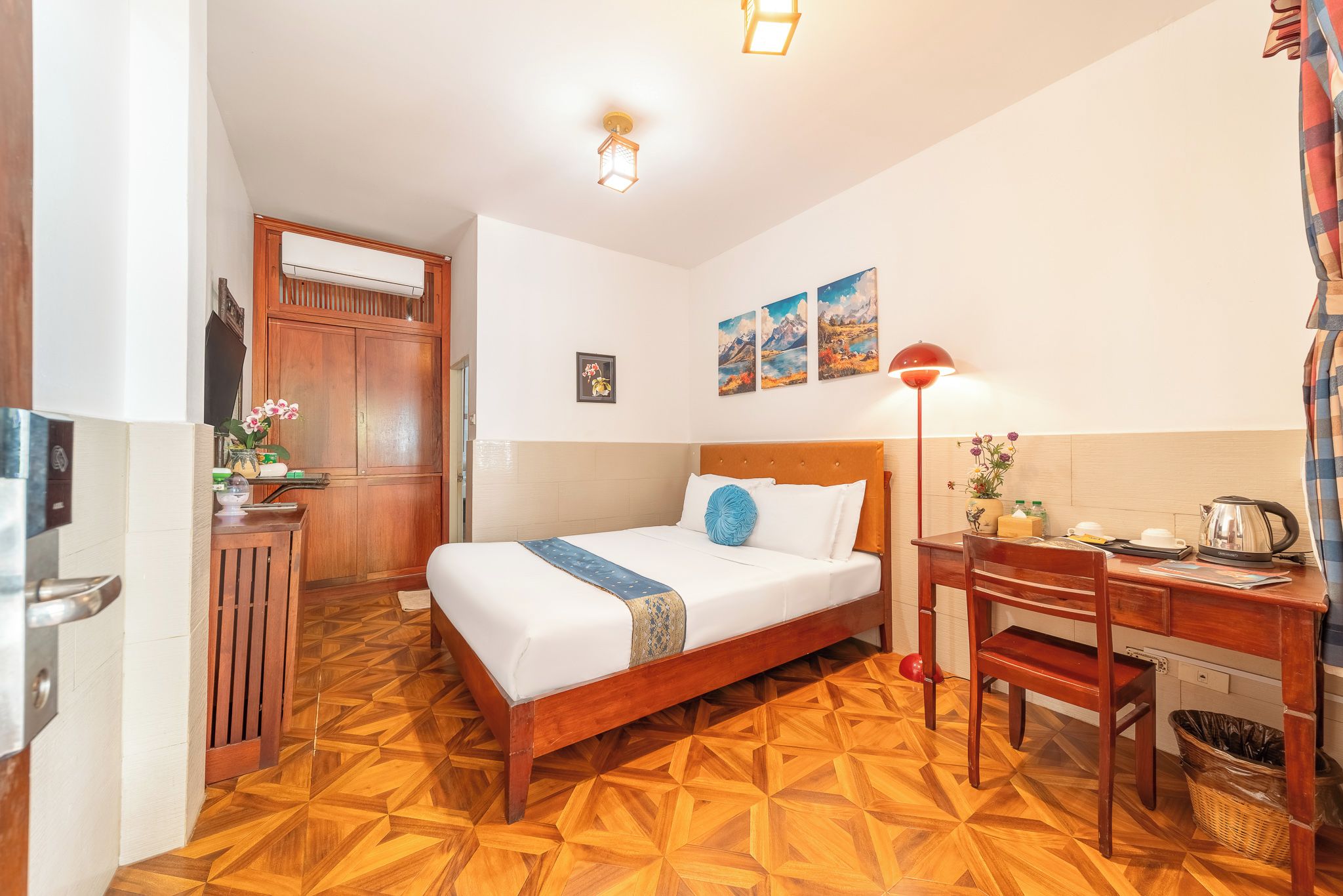 Habitación de hotel con piso de madera, cama con ropa de cama blanca y un cojín decorativo azul, escritorio con lámpara y aire acondicionado de pared.