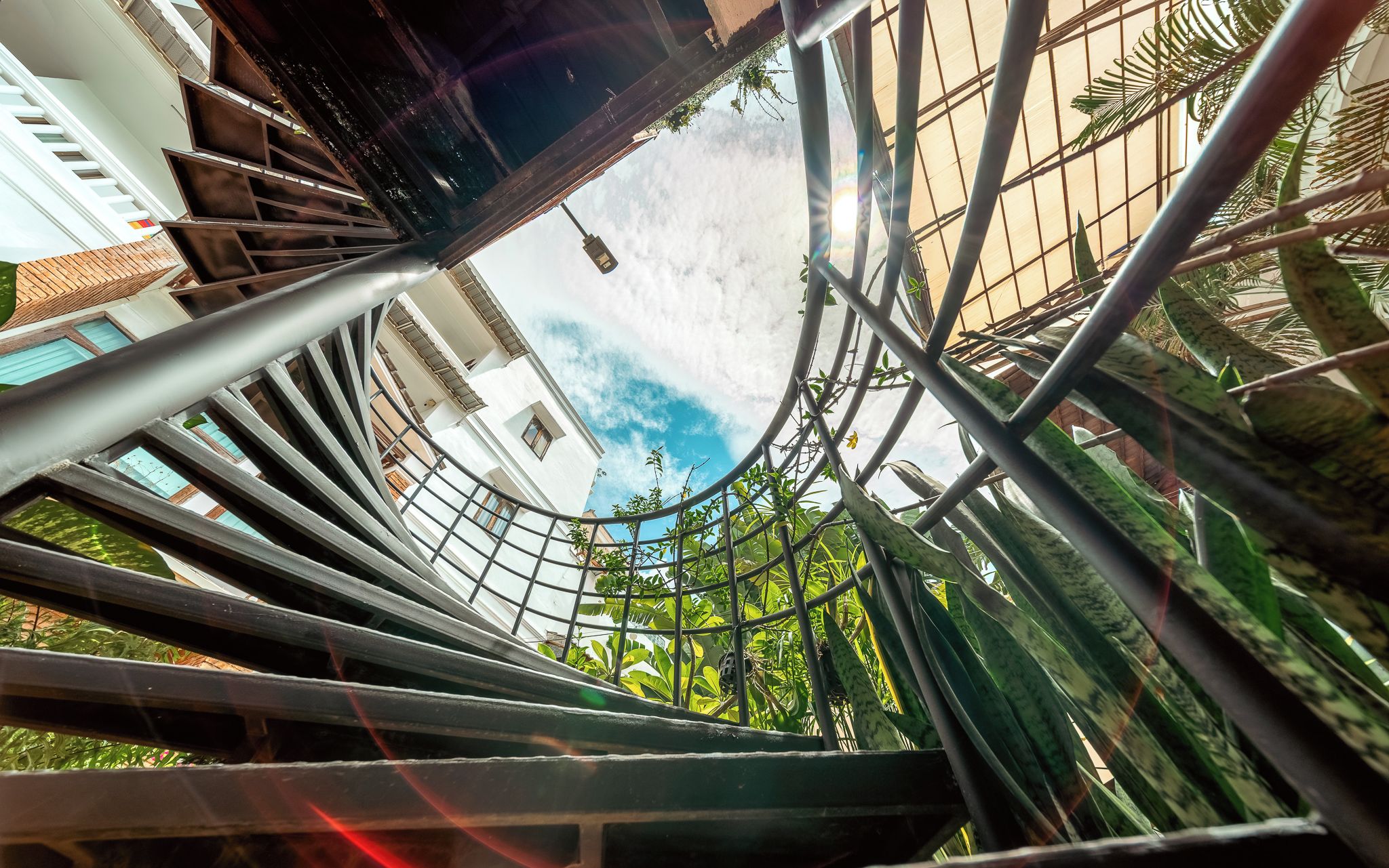 Escadaria em espiral com corrimãos de metal e vista para edifícios e vegetação ao fundo.