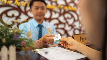 A person handing over a card to a receptionist at a reception desk with a floral arrangement and a decorative wall in the background.
